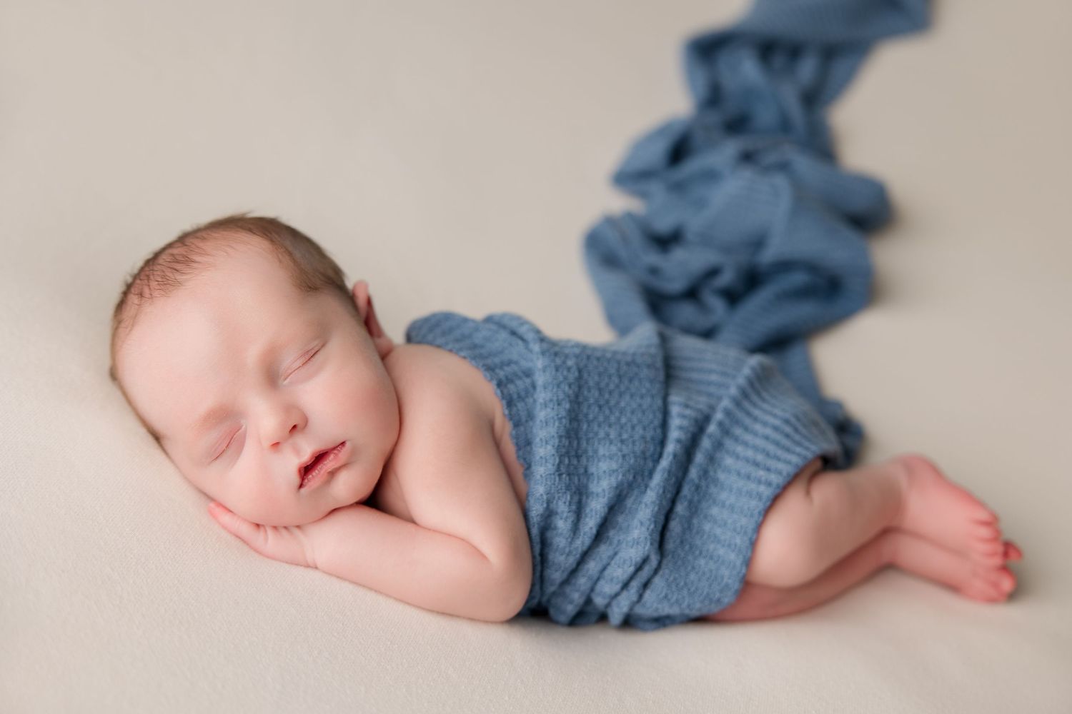 Sleeping newborn photographed in cool blue tones with soft fabric wrap against neutral beige background.