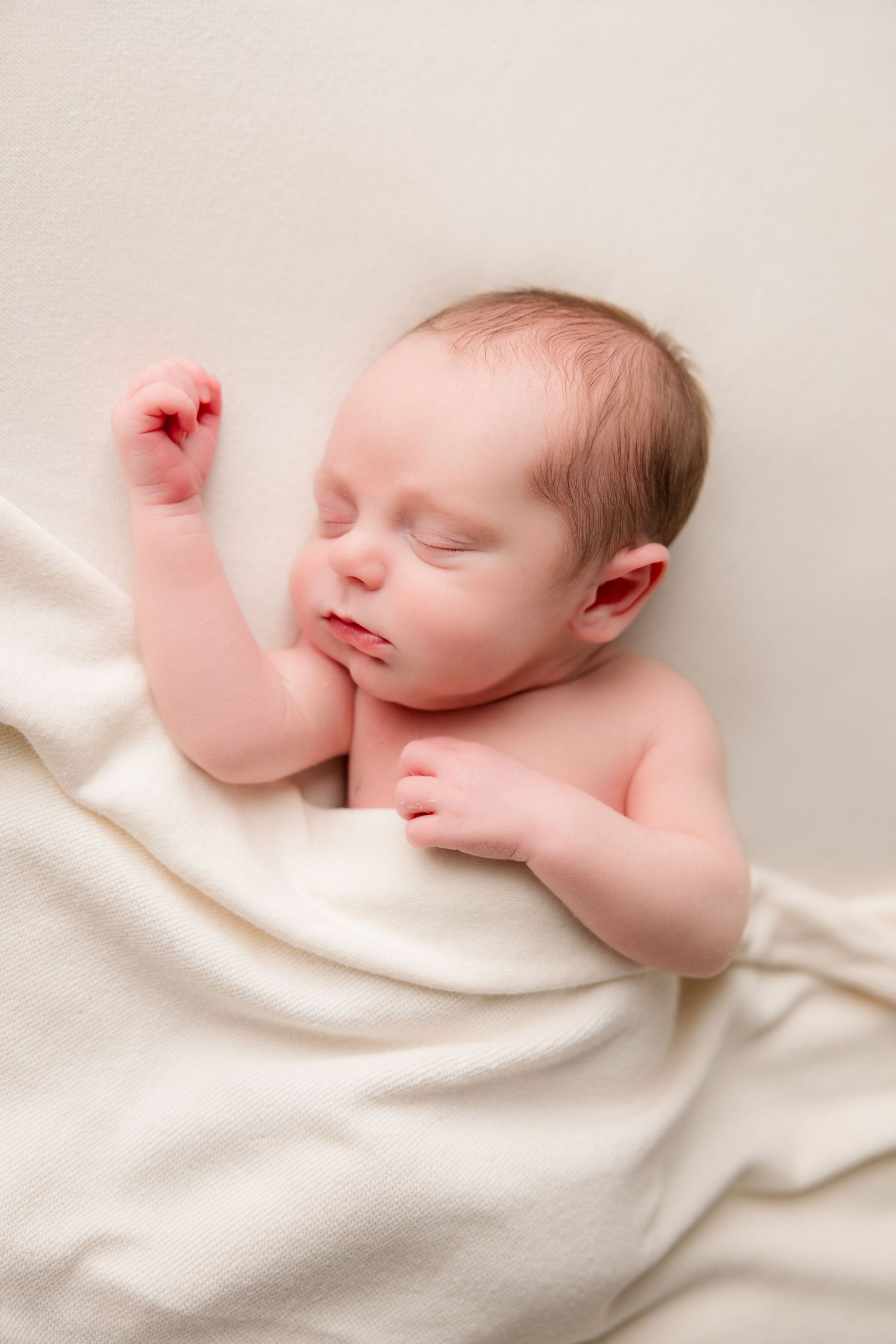 Peaceful sleeping newborn baby wrapped in white blanket on cream colored background.