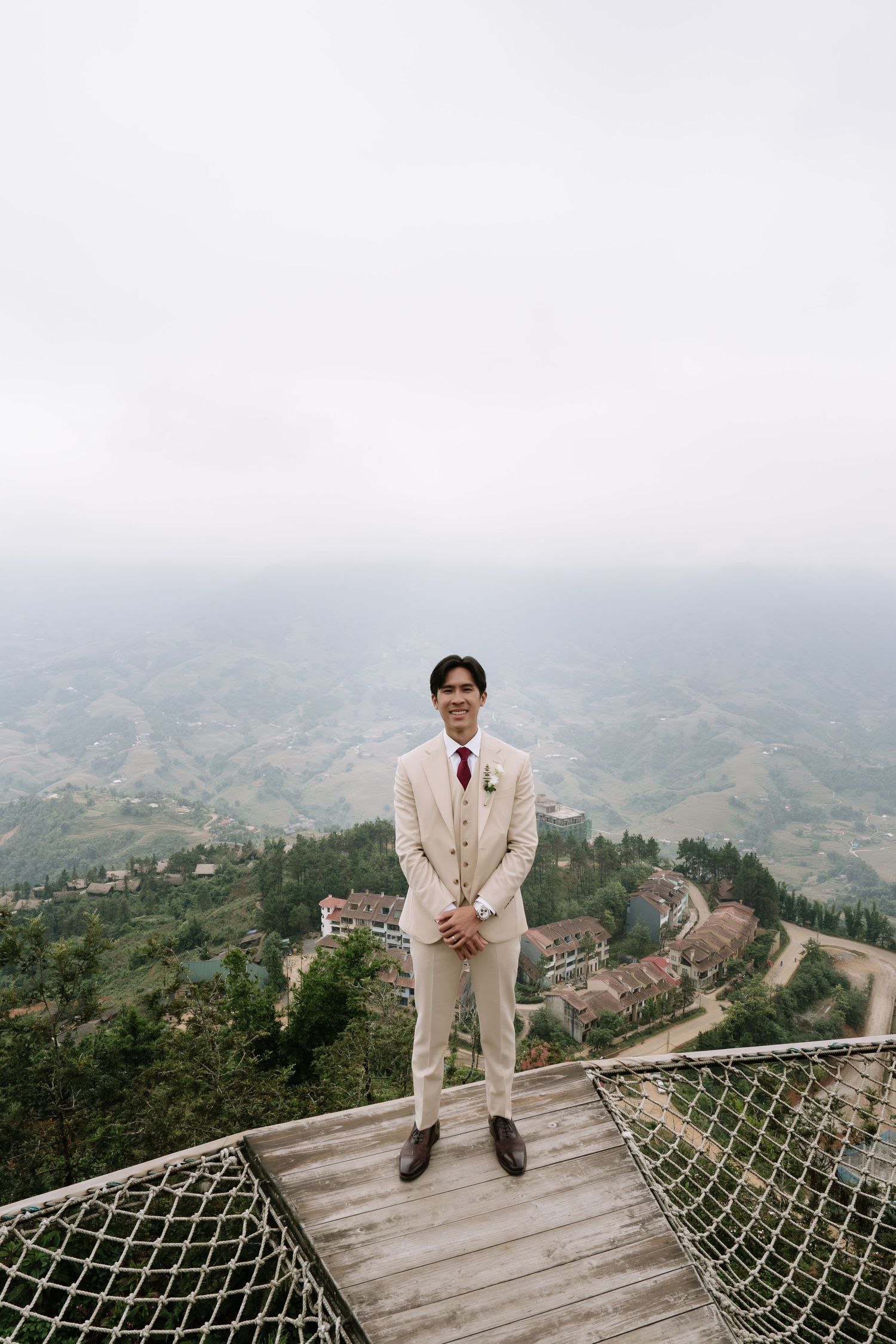 Dapper groom in cream suit stands on wooden mountain viewpoint overlooking lush green valley below.
