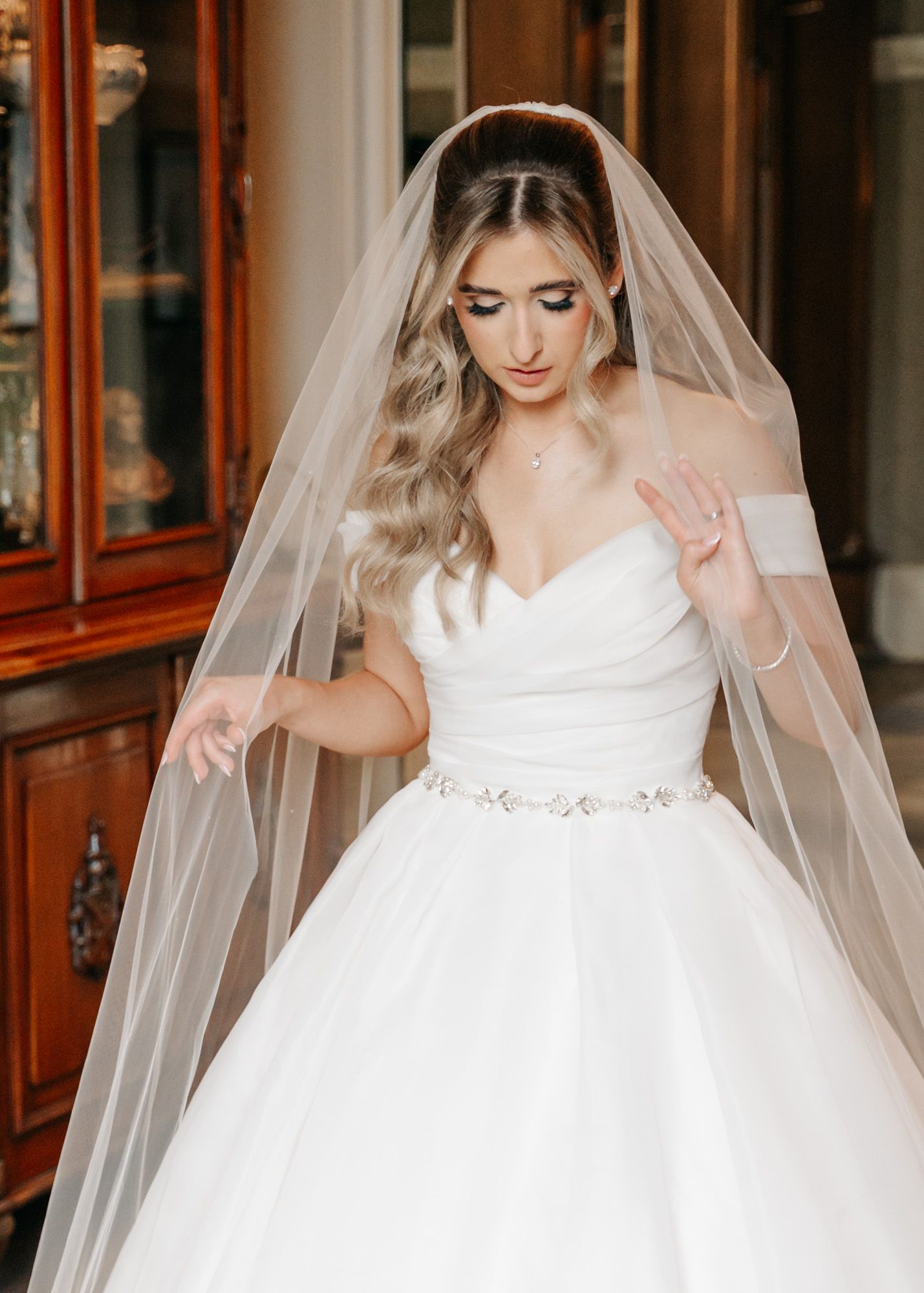 Bride in princess ballgown adjusts her flowing veil in elegant indoor setting.