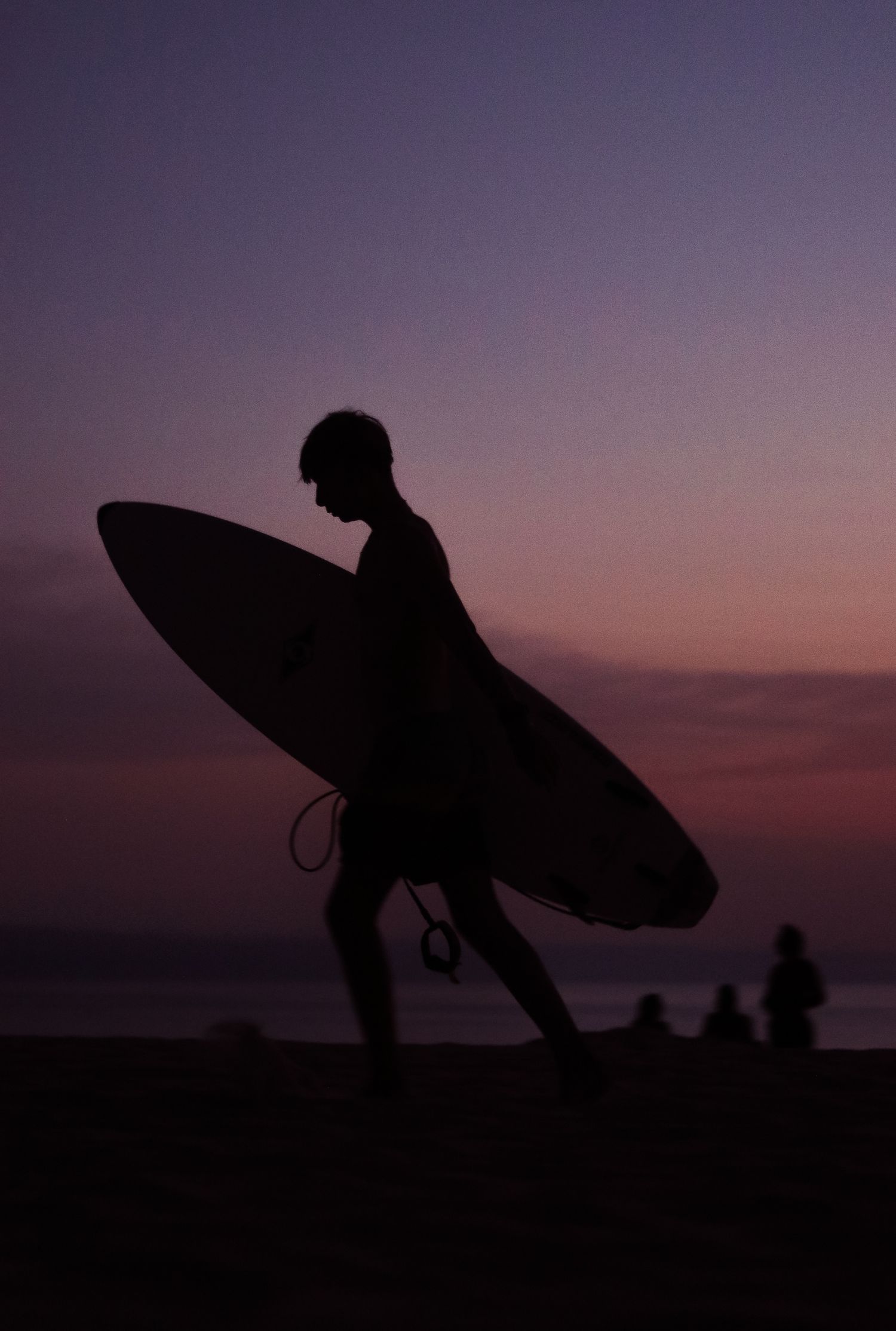 Silhouetted surfer carrying a surfboard walks along the beach against a dramatic purple sunset sky.