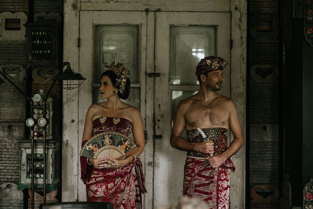 Wedding couple in traditional red Balinese attire posed against rustic wooden backdrop.