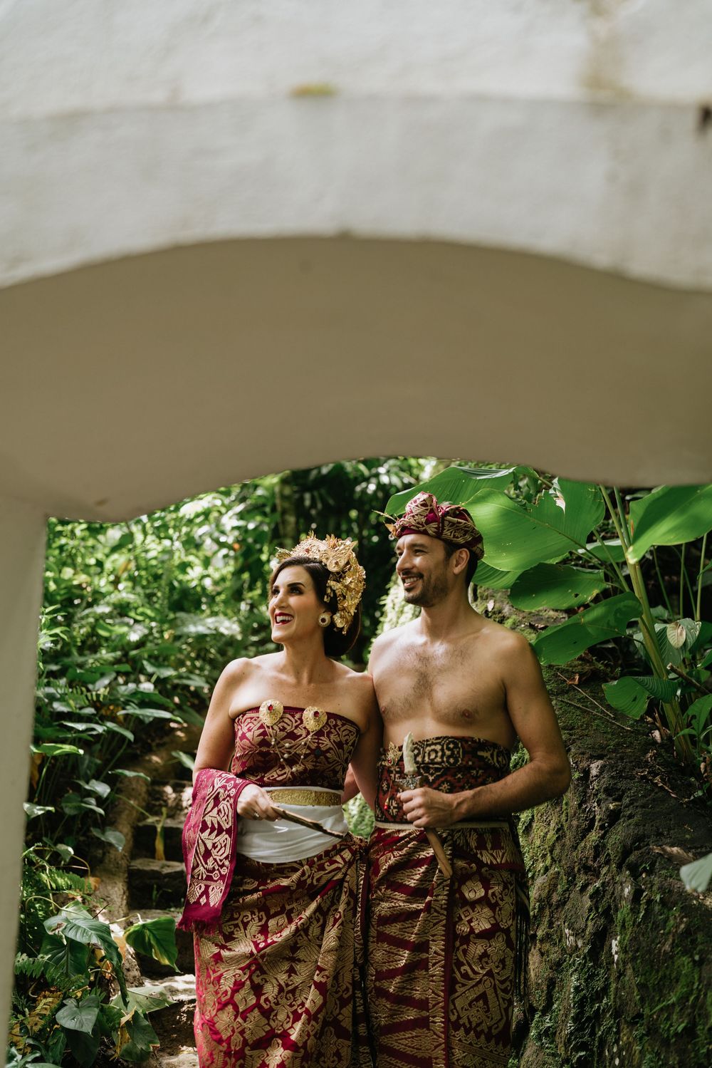 A couple wearing traditional Balinese attire share a joyful moment together in a lush tropical garden setting.