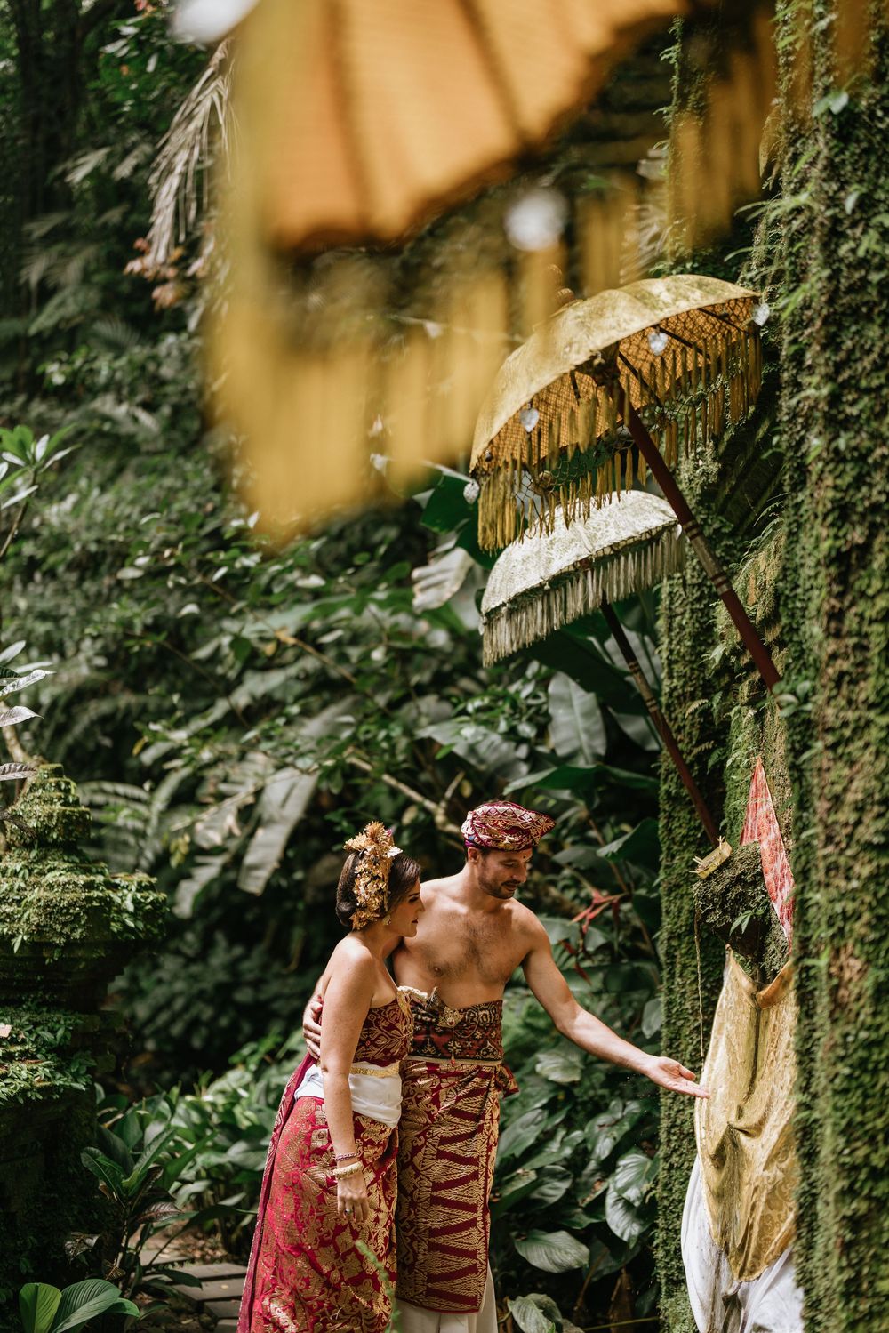 A couple in traditional Balinese dress pose beneath decorative umbrellas in a lush garden setting.