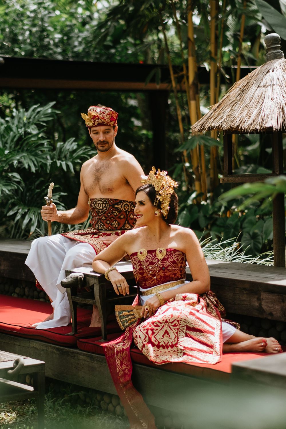 Traditional Balinese wedding couple seated on wooden platform surrounded by tropical foliage.