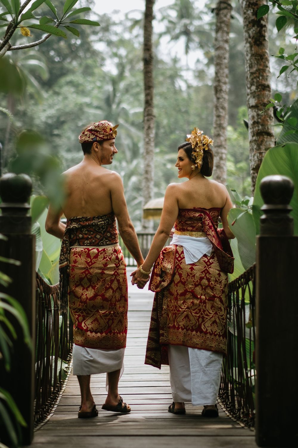 Two people in traditional Indonesian sarongs sit together on a wooden bridge surrounded by tropical foliage.