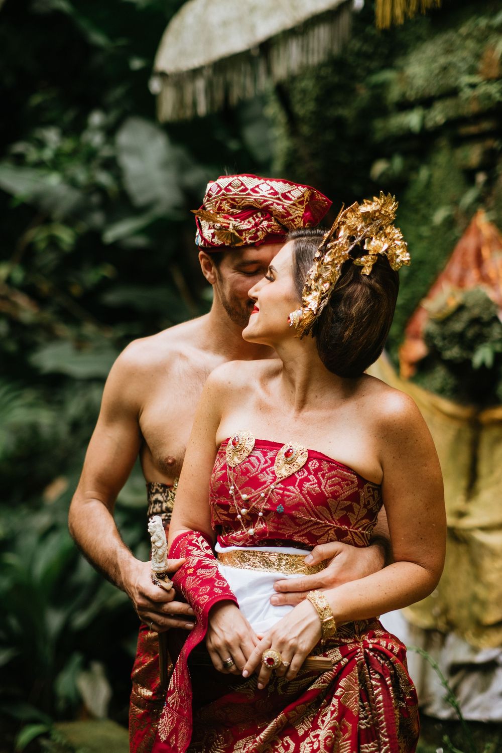 Intimate moment captured between couple wearing red and gold Balinese wedding attire.