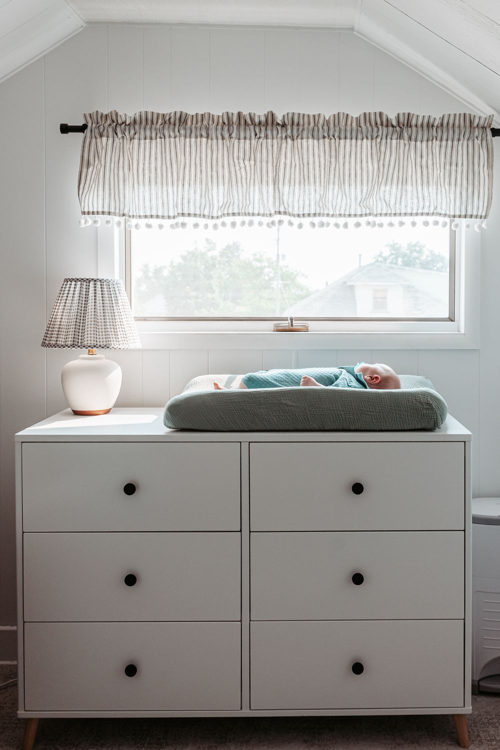A white dresser with changing pad sits beneath a window with striped valance and table lamp.