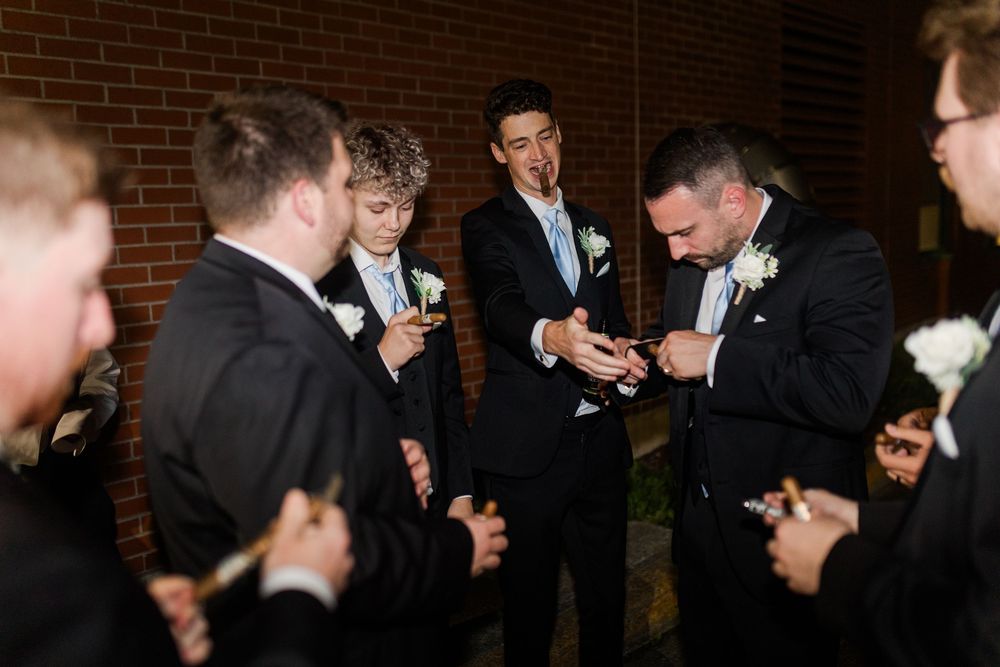 Wedding party members check their phones while standing against a brick wall at a formal event.
