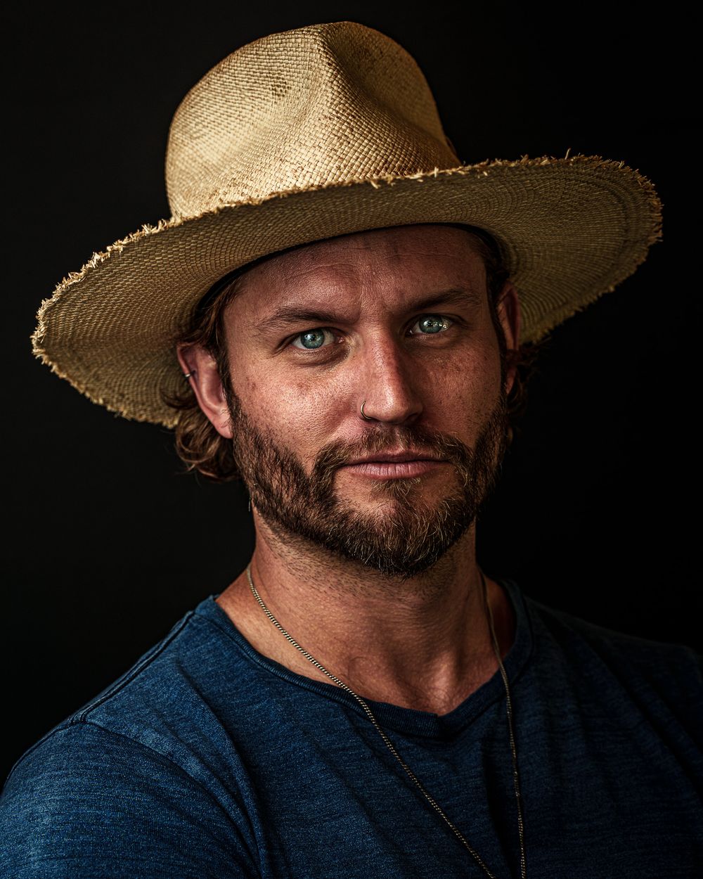 Ryan Mcgrath wearing a custom straw hat and denim shirt poses against a dark background in dramatic lighting.