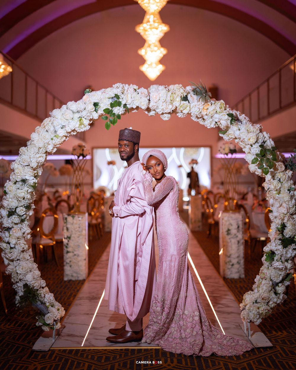 Romantic wedding moment captured under a floral arch with soft pink lighting and white rose decorations.
