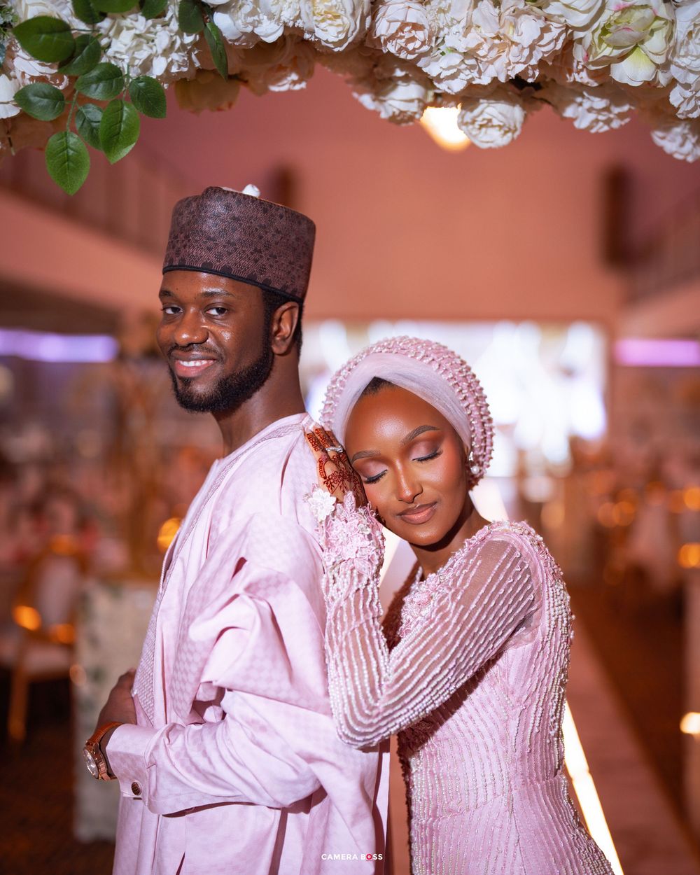 Romantic wedding couple poses in coordinated white traditional outfits during indoor reception photography session.