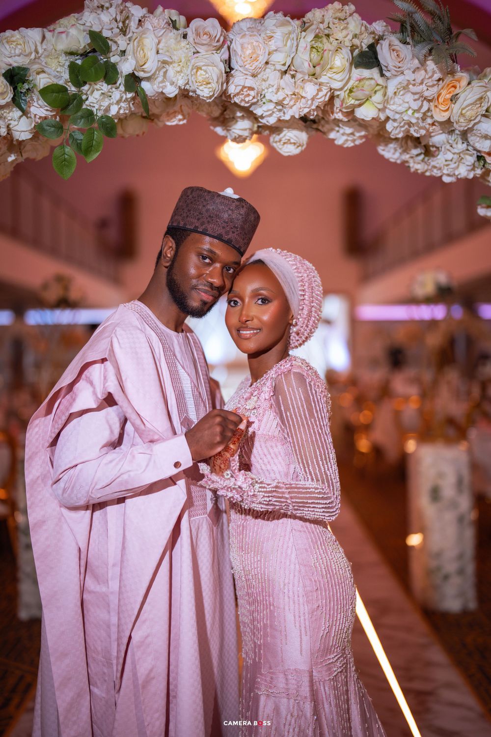 A wedding celebration featuring traditional pink and white Nigerian attire with floral decor and warm lighting.