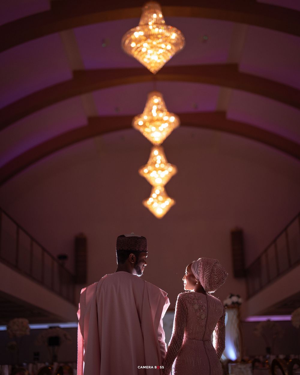 A couple in traditional dress stand beneath illuminated chandeliers creating a romantic silhouette in an elegant venue.