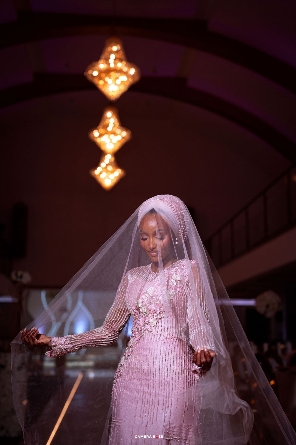 A bride in a beaded gown and veil poses dramatically under warm lighting.