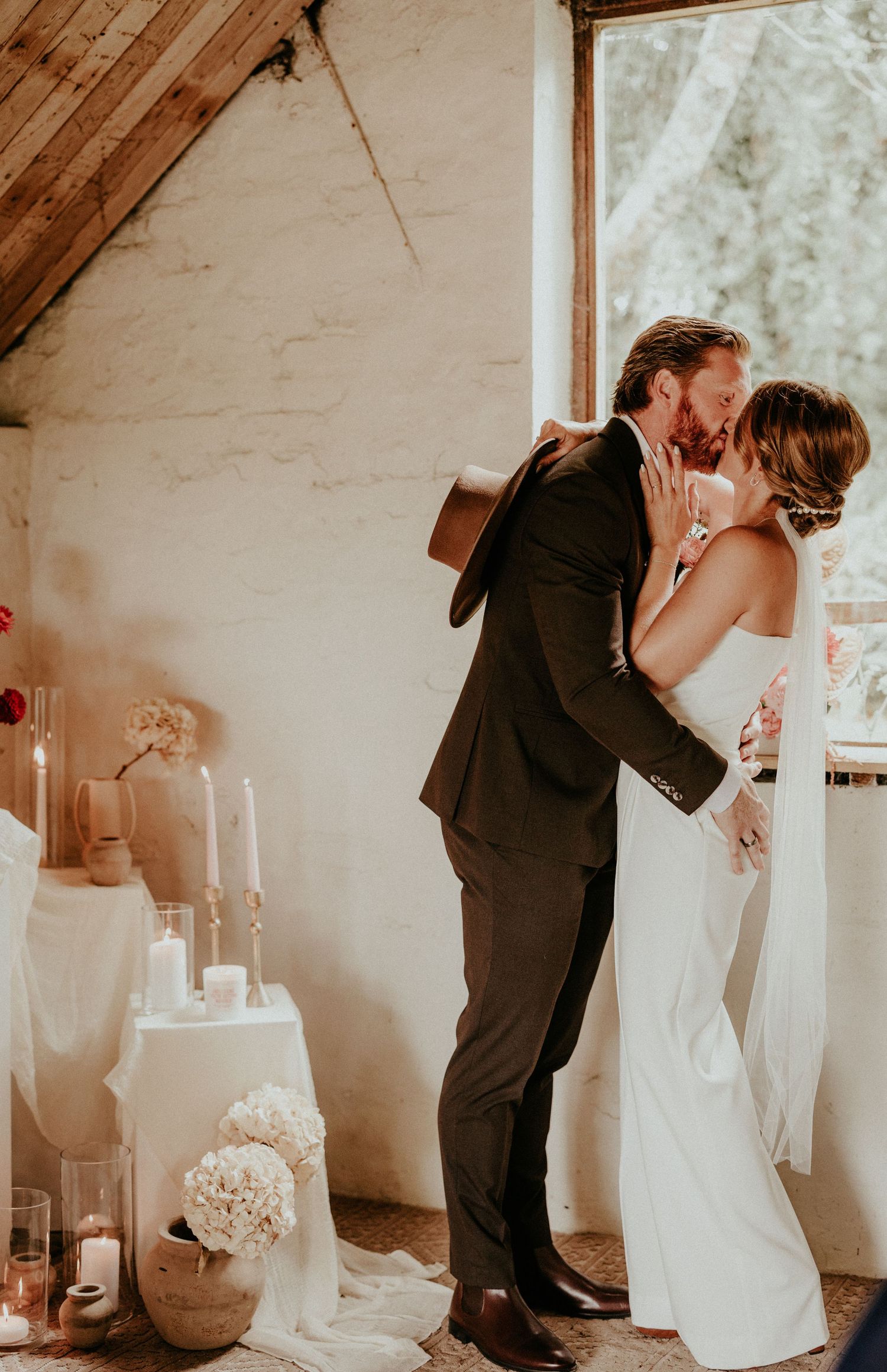 A romantic first kiss between newlyweds at an intimate wedding ceremony in a rustic white interior space.