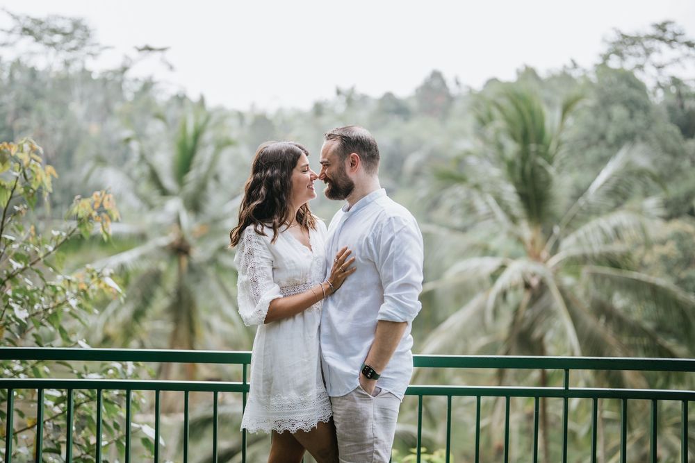 A couple in white outfits share an intimate moment on a balcony overlooking a tropical palm tree landscape.