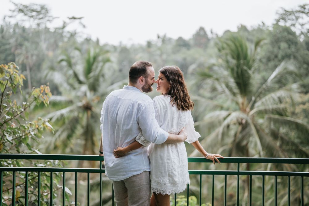 A romantic couple embraces on a balcony with lush palm trees and greenery in the background.