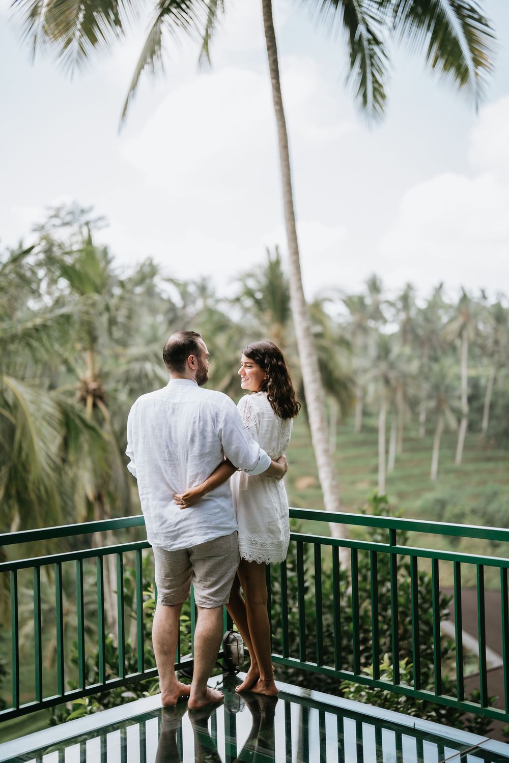 Two people in white clothing share a tender moment on a balcony surrounded by tropical palm trees.