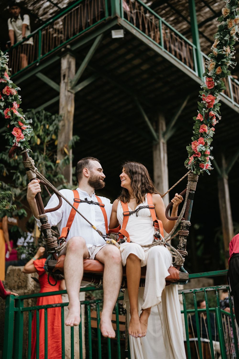 Couple sits playfully on wooden railings decorated with floral arrangements at a tropical venue.