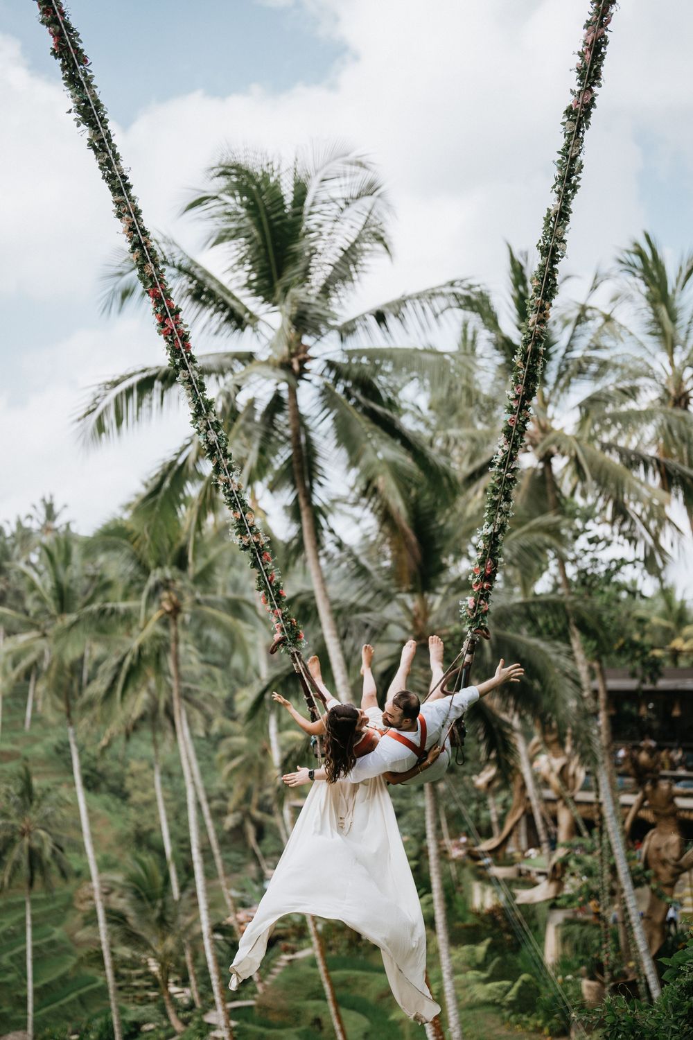 A couple swings together on a flower-decorated swing with palm trees in the background.