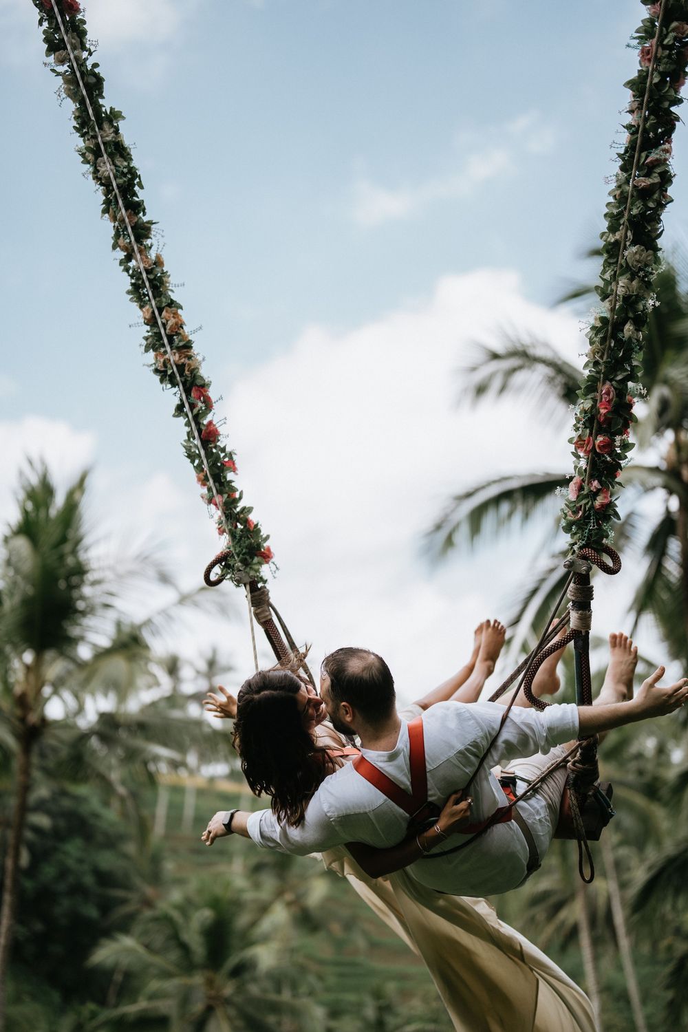 Two people swing playfully on a floral-decorated rope swing against a tropical palm tree backdrop.