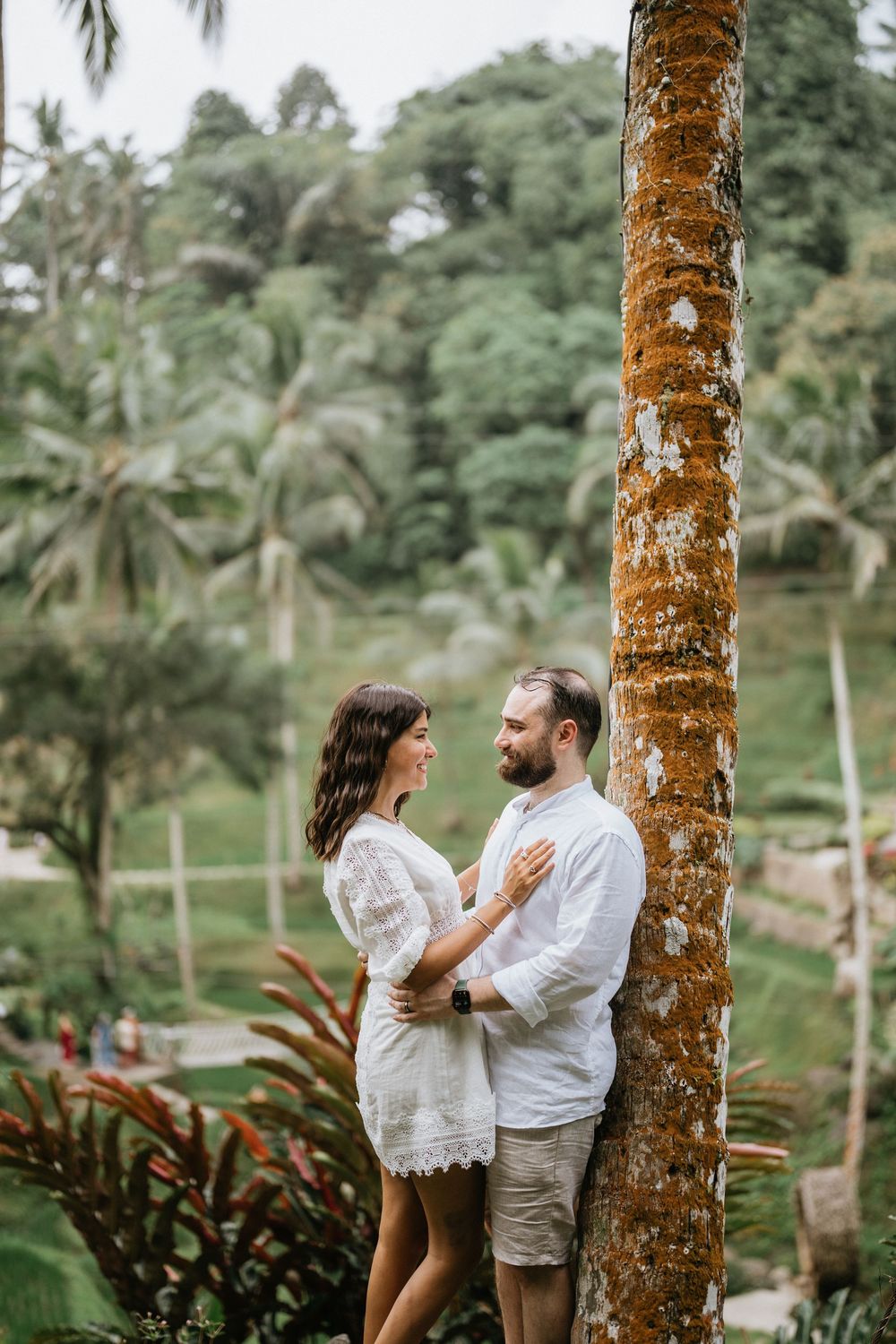Couple shares a playful moment near a palm tree trunk in a lush tropical setting.