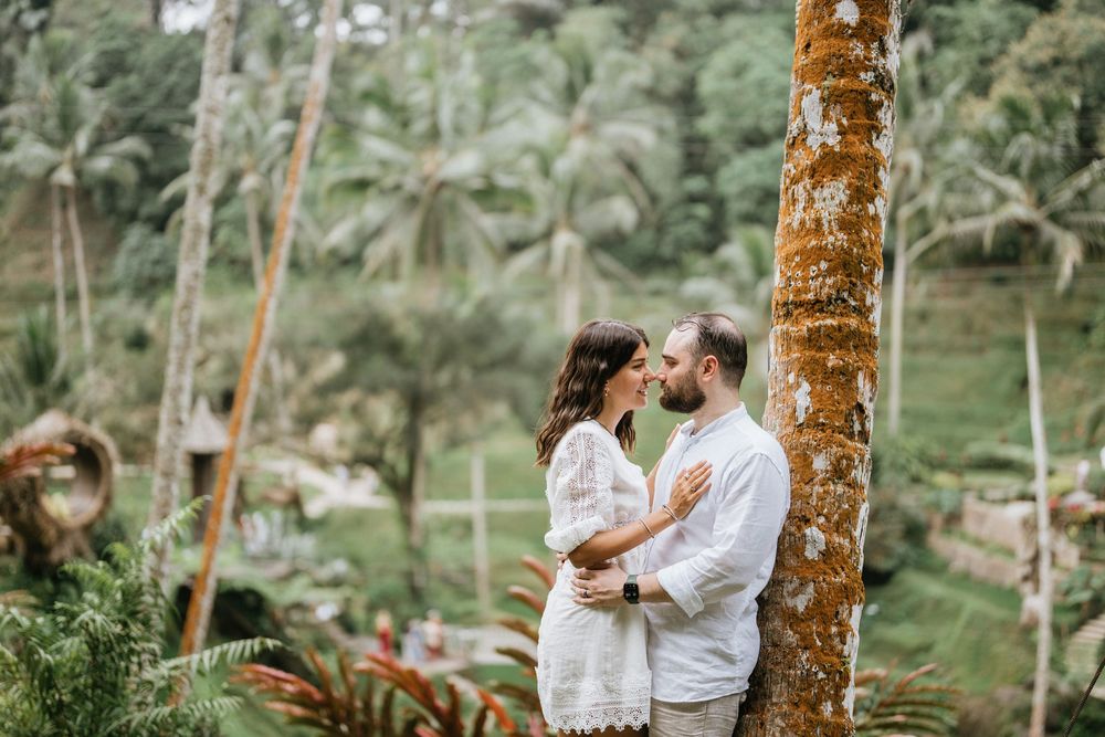 Two people in white outfits embrace near palm trees in a tropical garden setting.