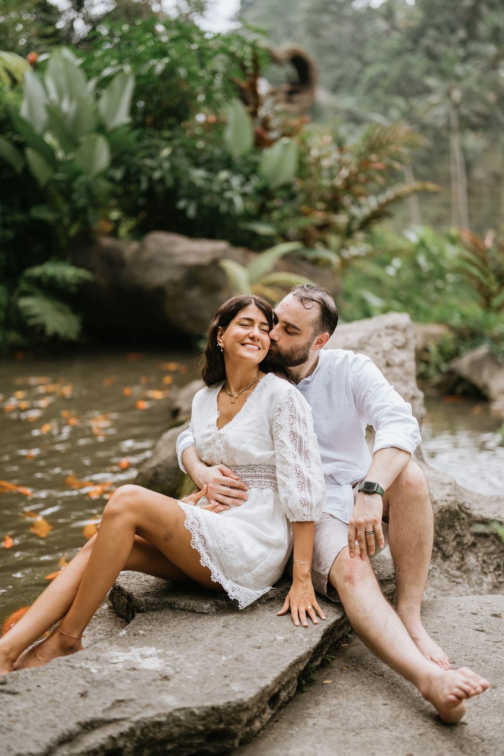 Couple in white clothing shares intimate moments by a stream in a tropical garden.