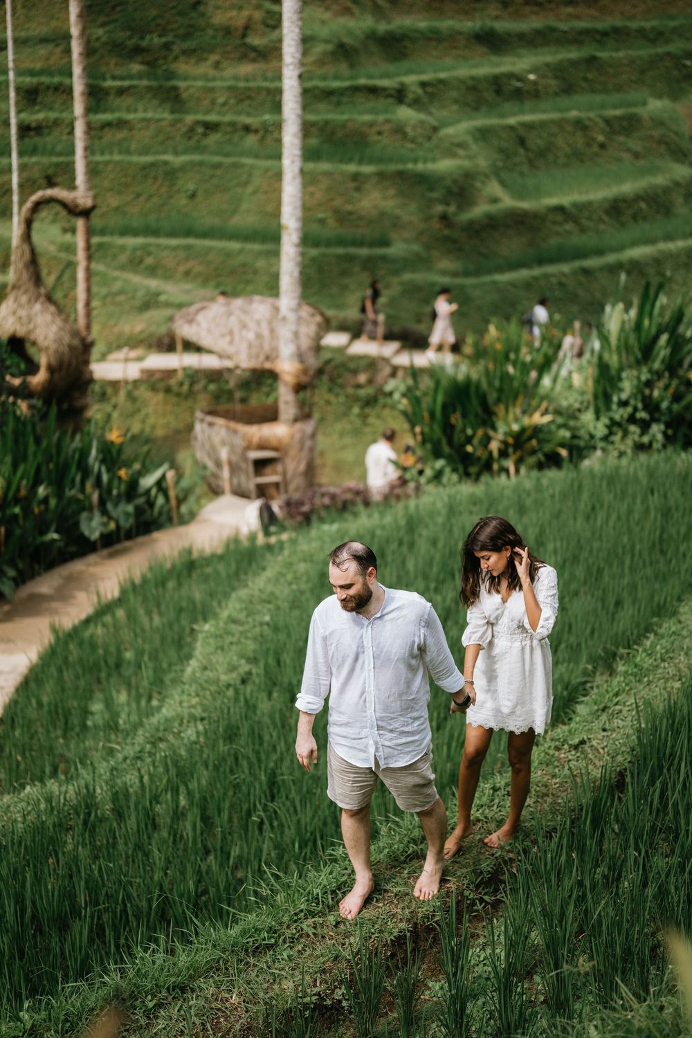 Two people in light clothing walk through terraced rice fields surrounded by tropical greenery.