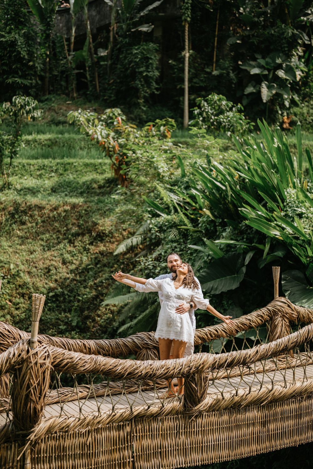 Two people posing playfully on a traditional woven bamboo bridge in a tropical setting.