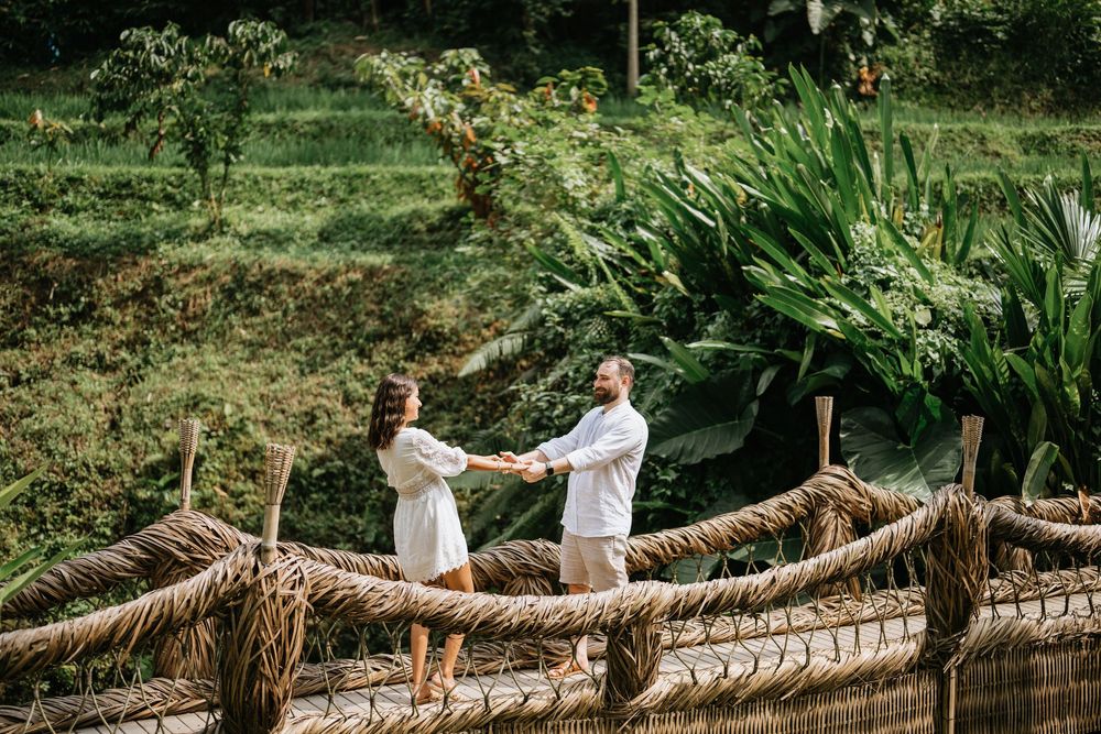 A couple dressed in white walking across a handcrafted bamboo bridge in a lush landscape.