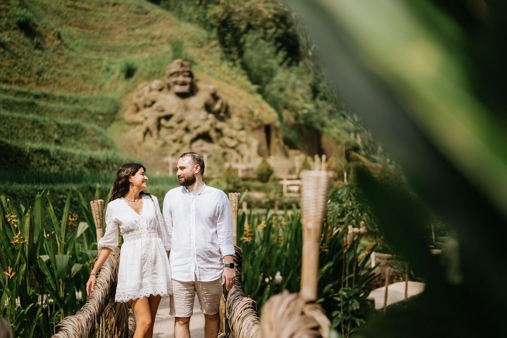 Two people in white outfits standing on a path surrounded by tropical greenery.