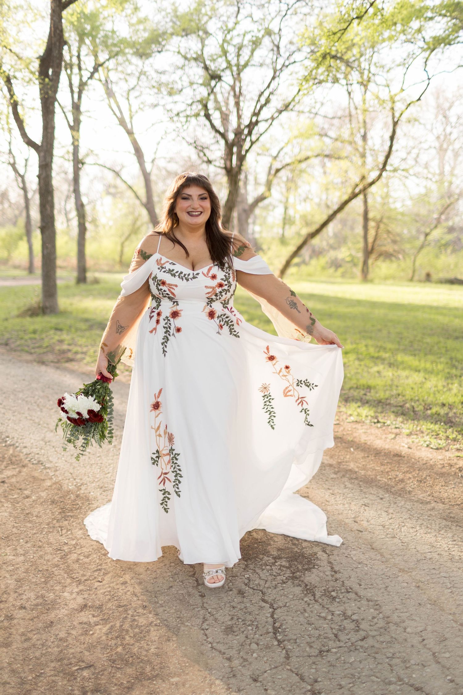 Bride in white floral embroidered off-shoulder gown holding bouquet, walking on park path among trees.