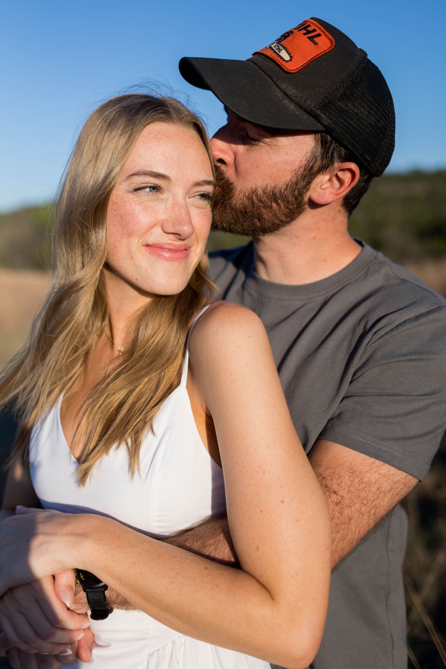 A man in a black cap kisses a smiling blonde woman in a white dress outdoors in golden light.
