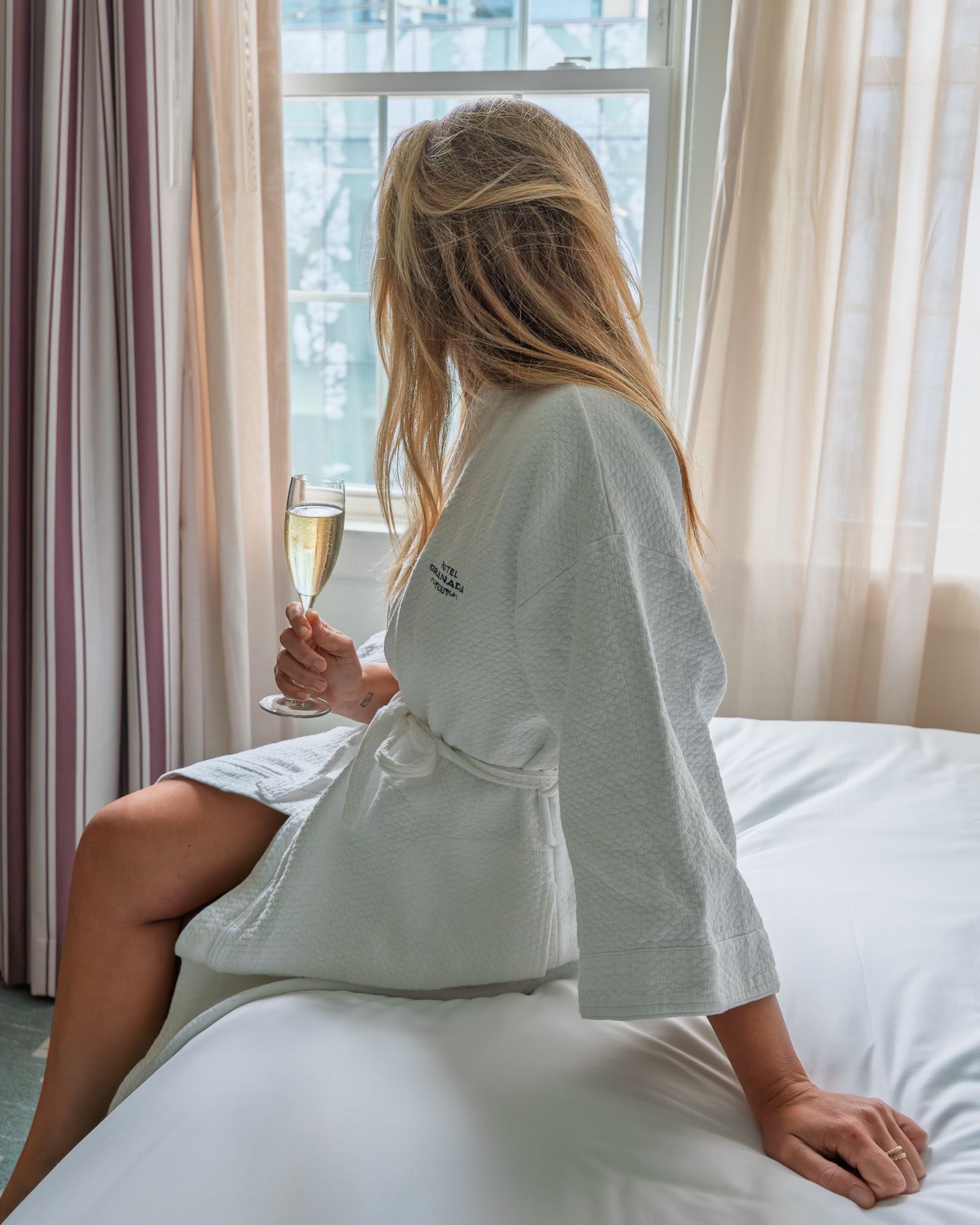 Blonde woman in white hotel robe sitting on bed holding champagne glass by window.