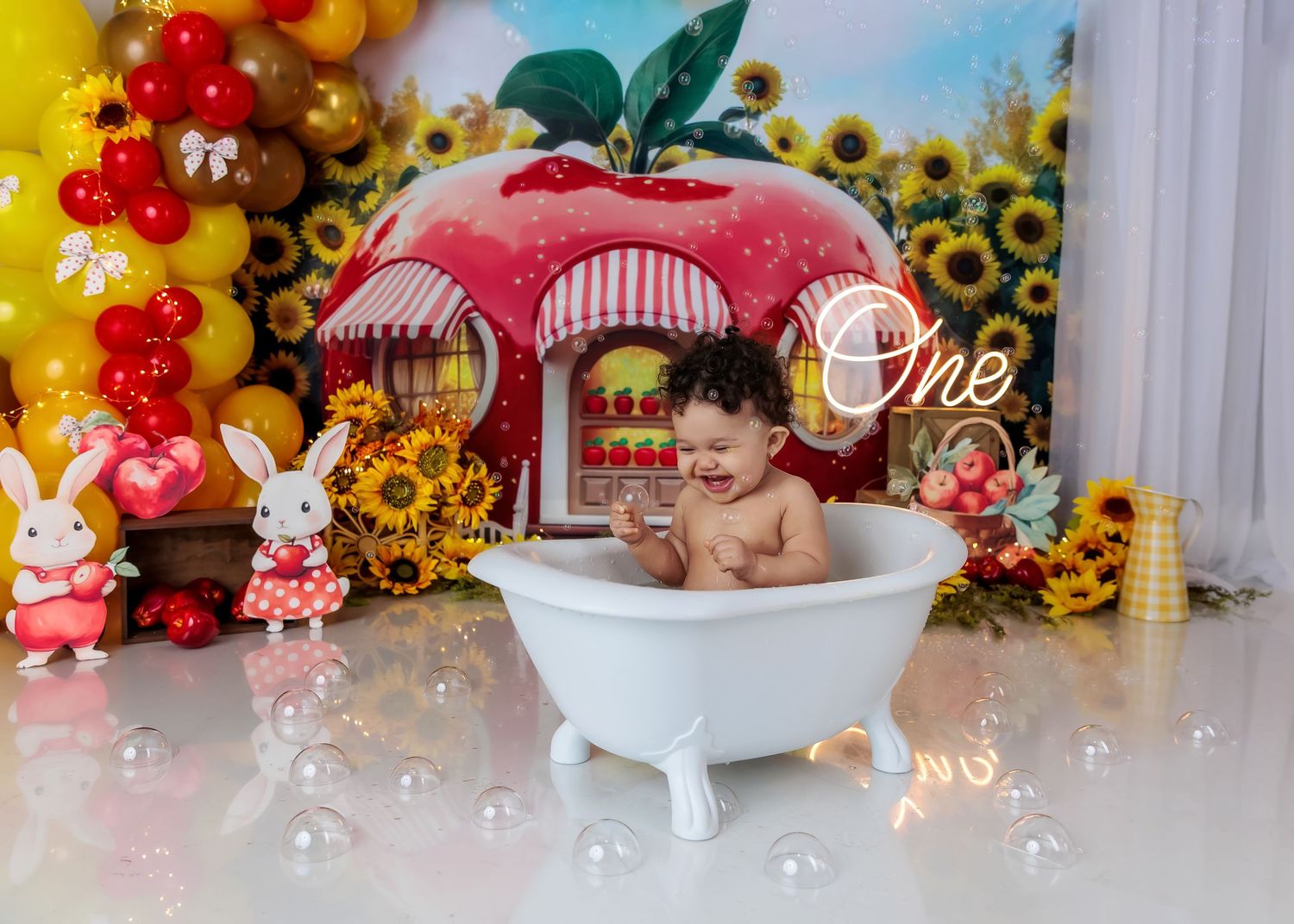 Baby sitting in a white bathtub during a Snow White themed first birthday cake smash with apple backdrop.