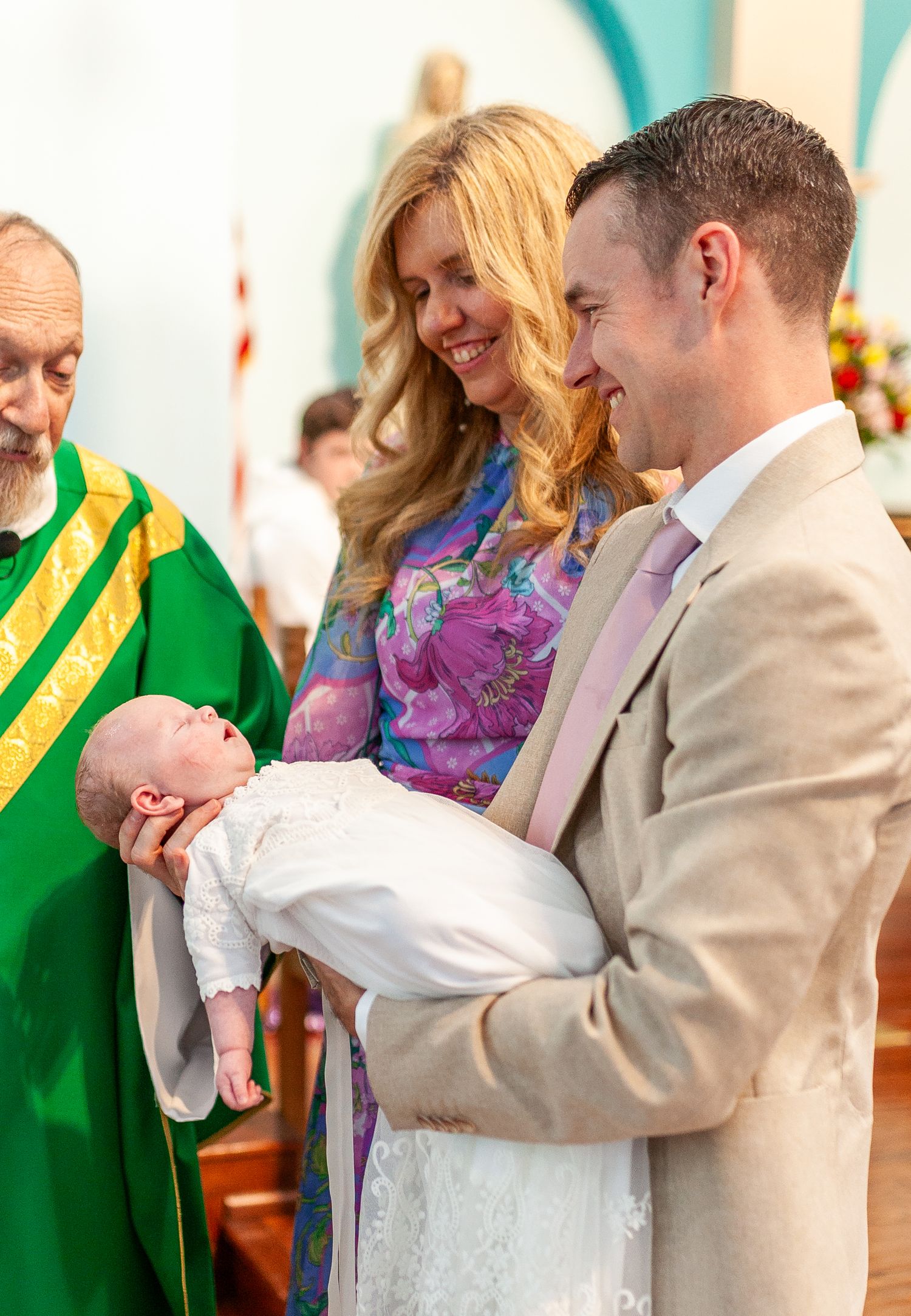 Couple smiling at newborn baby during baptism ceremony with priest in green vestments.