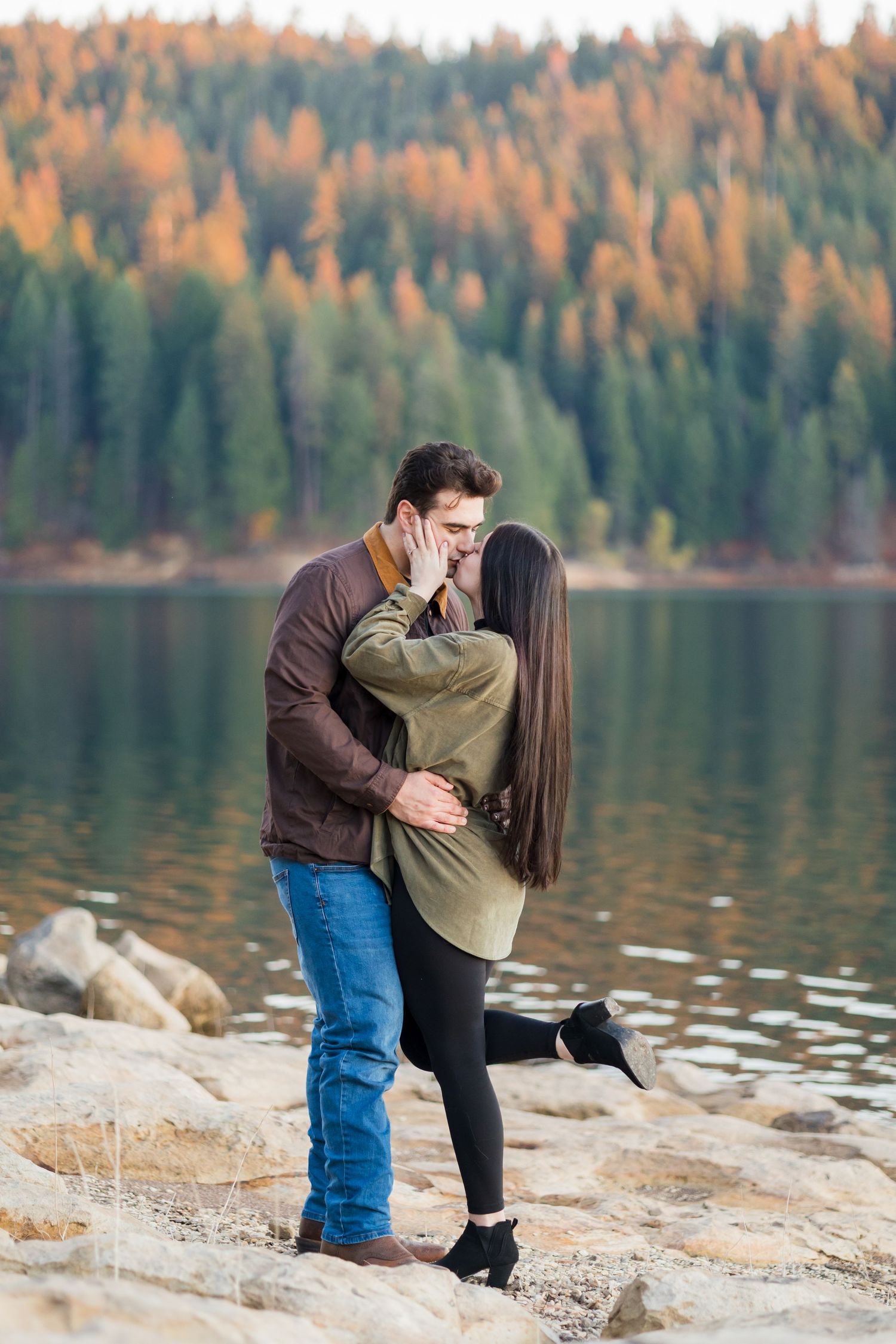 Couple sharing a romantic kiss by a mountain lake in autumn, surrounded by colorful fall foliage reflecting on the water.
