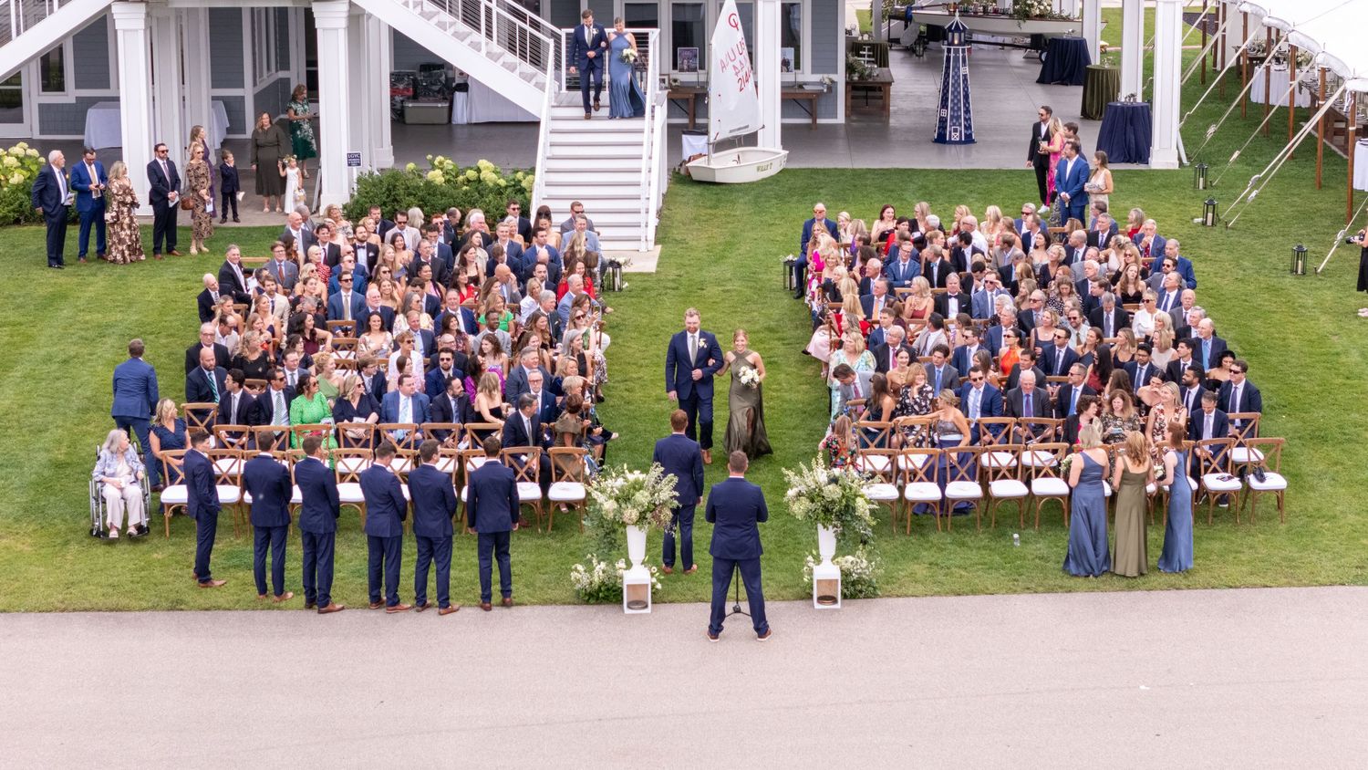 Aerial view of a large outdoor wedding ceremony with guests seated on a green lawn beside a white building.
