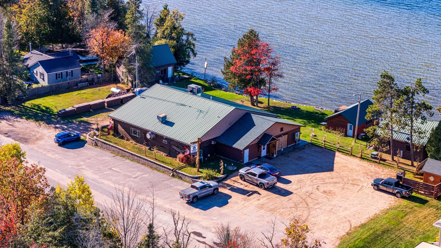 Aerial view of a lakeside property with a green-roofed building, parking area, and colorful autumn trees along the waterfront.