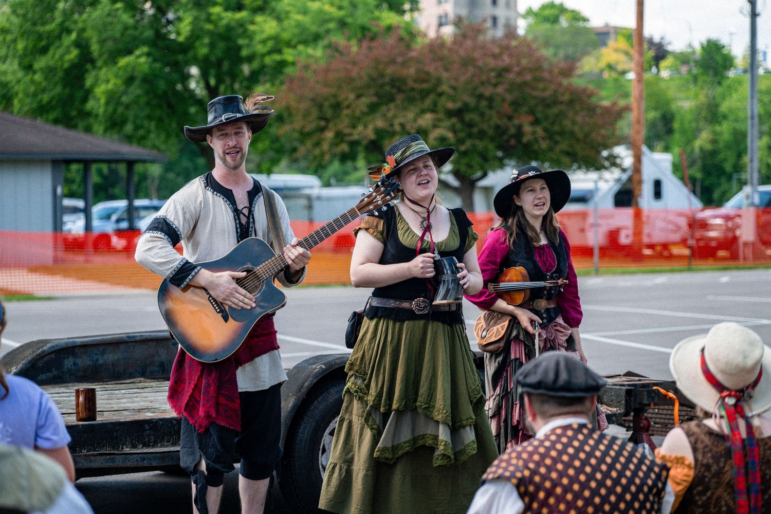 Three Renaissance faire performers in period costumes play guitar and violin outdoors at a festival.