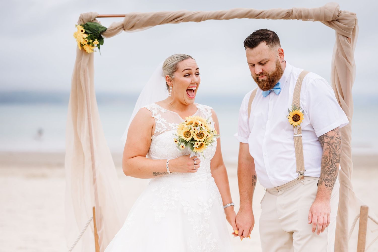 Bride and groom laughing holding hands under rustic wooden arch on beach with sunflower bouquet and boutonniere. Beach wedding UK