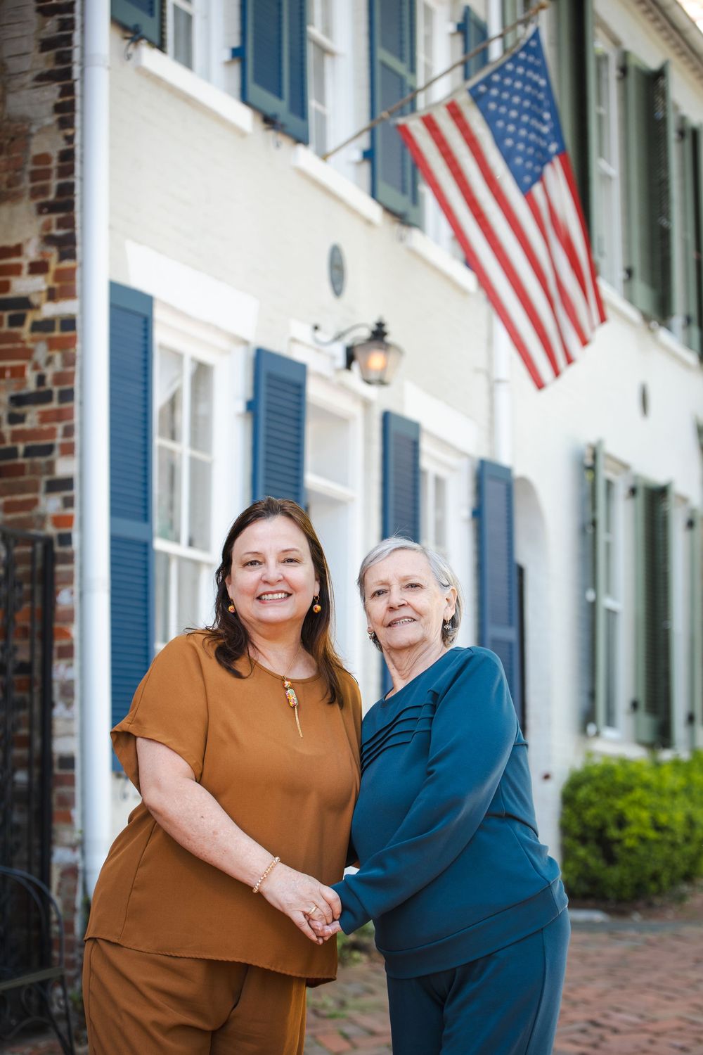 Two smiling women standing together outdoors in front of a white building with an American flag.
