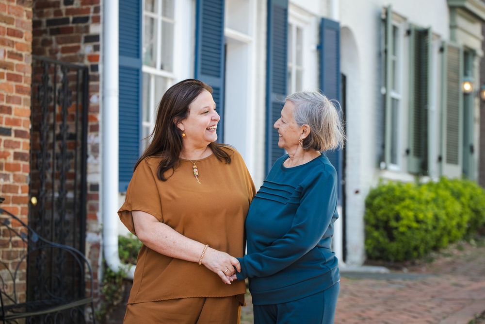 Two women smiling and holding hands outside a historic brick building with blue shutters.