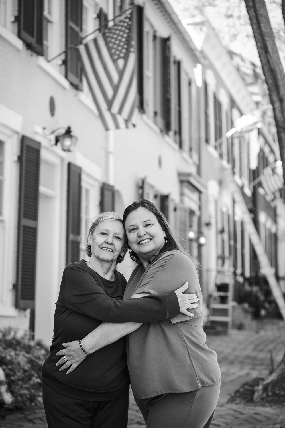 Two women embracing and smiling outdoors on a cobblestone street with American flags on buildings.