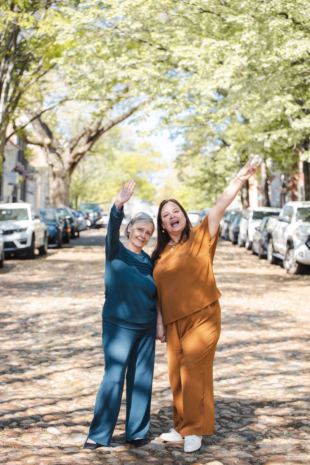 Two women in colorful outfits joyfully raising their arms on a tree-lined cobblestone street on a sunny day.
