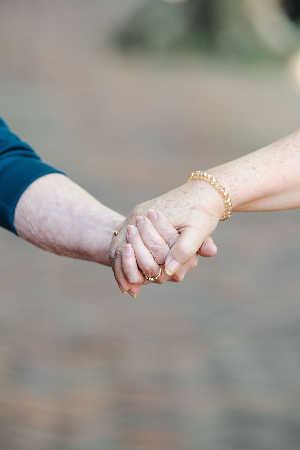 Elderly couple holding hands tenderly, woman wearing gold bracelet and ring, showcasing love and togetherness outdoors.