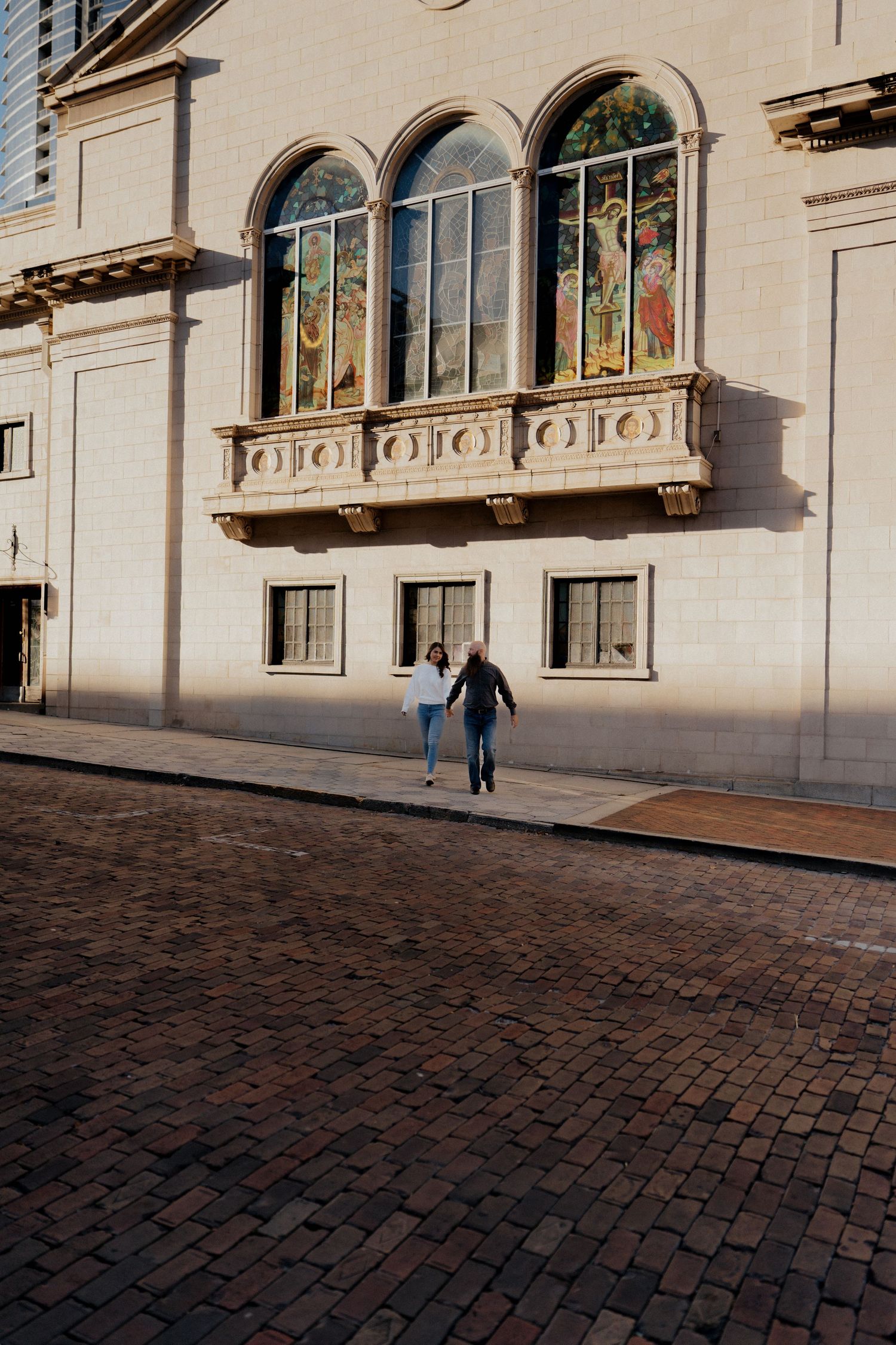 Couple walking hand in hand on cobblestone street beside ornate building with arched stained glass windows.