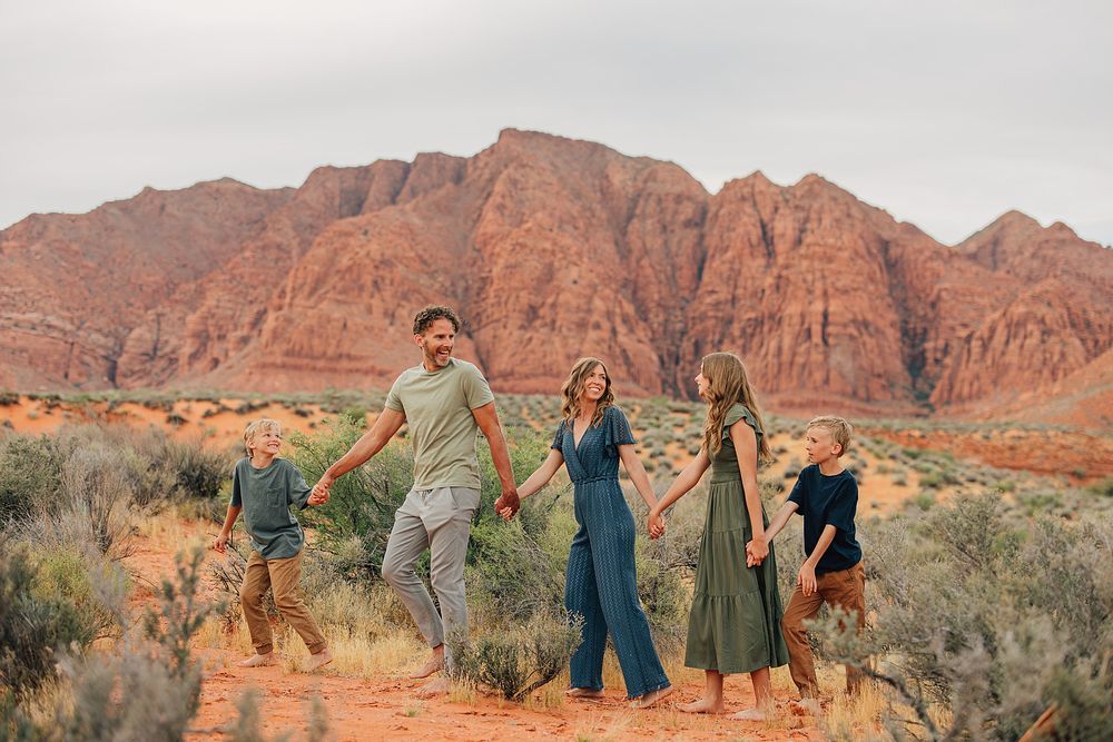 Family of five holding hands walking in red rock desert landscape with dramatic sandstone cliffs backdrop.